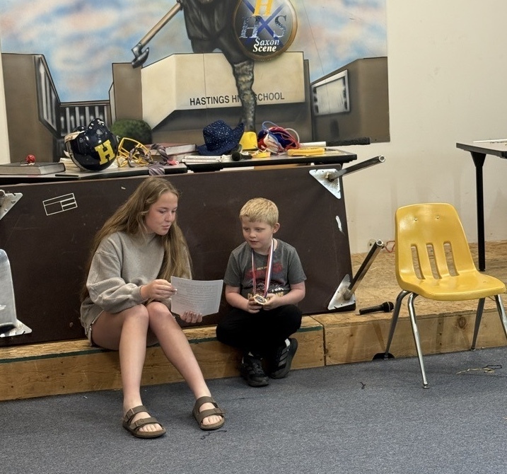 A high school student sits next to a young student and shows him writing on a piece of paper