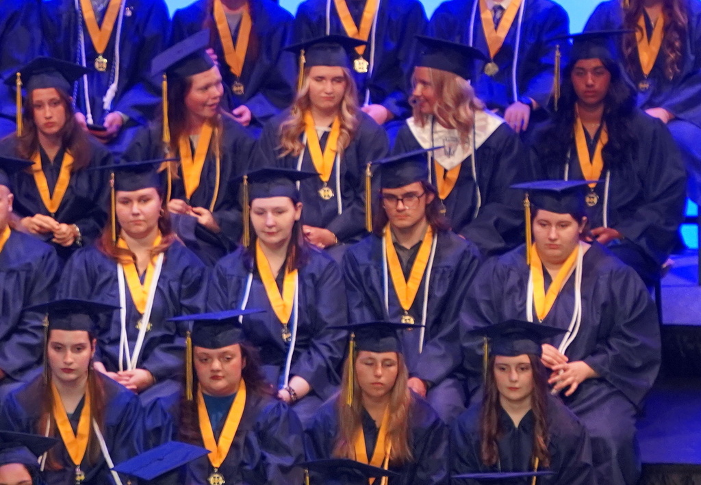 Students in graduation gowns sit in bleachers as people speak at a podium