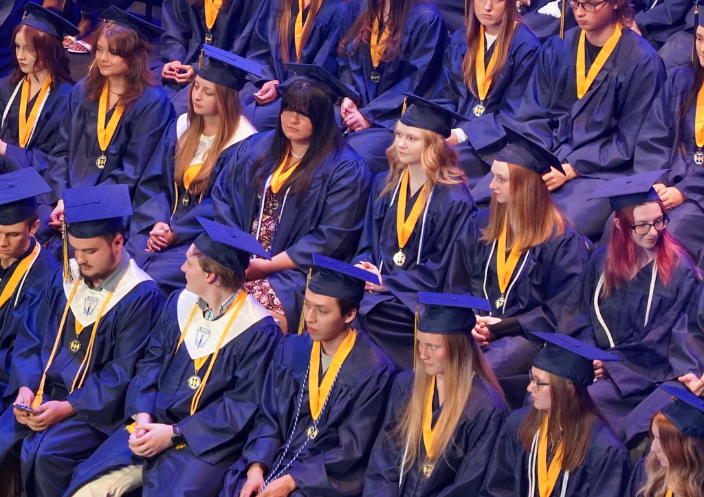 Students in graduation gowns sit in bleachers as people speak at a podium
