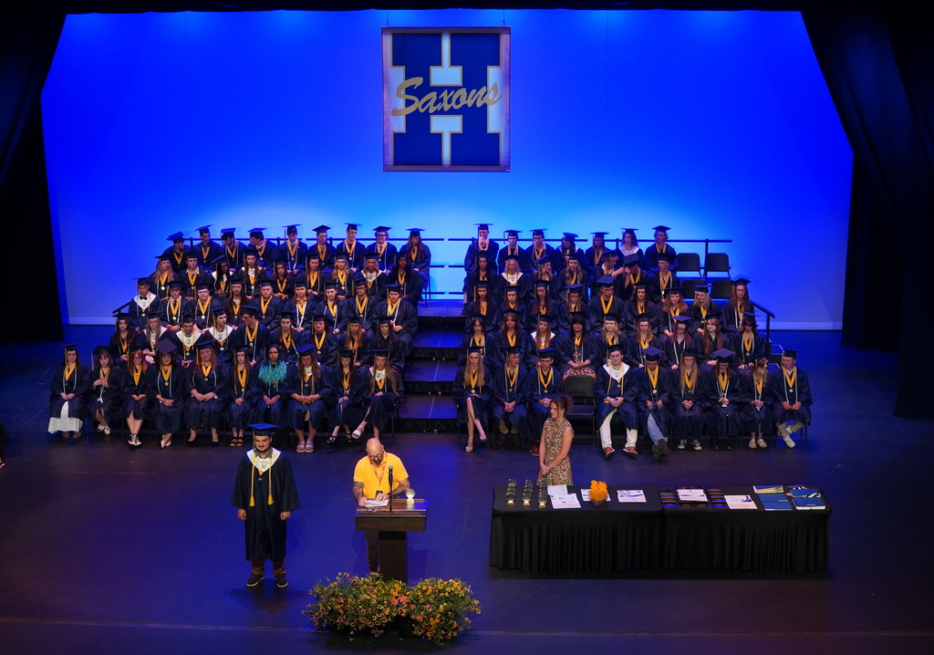 Students in graduation gowns sit in bleachers as people speak at a podium