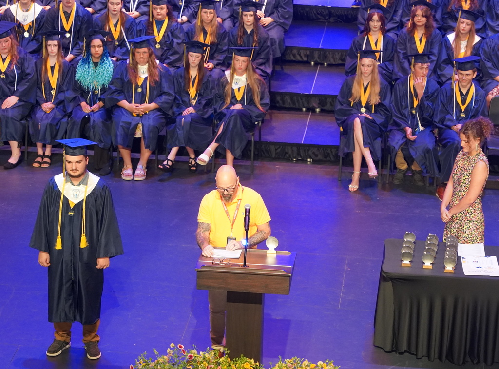 Students in graduation gowns sit in bleachers as people speak at a podium