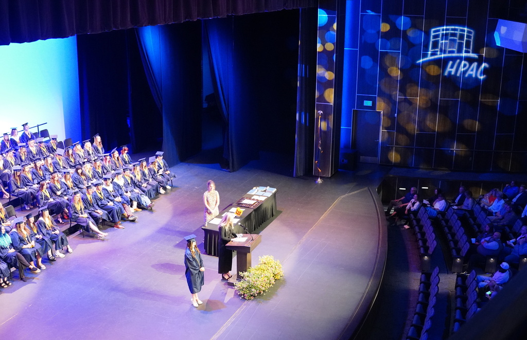 Students in graduation gowns sit in bleachers as people speak at a podium