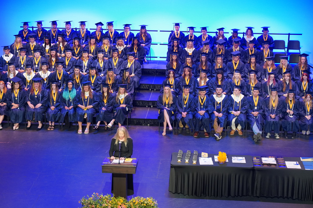 Students in graduation gowns sit in bleachers as people speak at a podium
