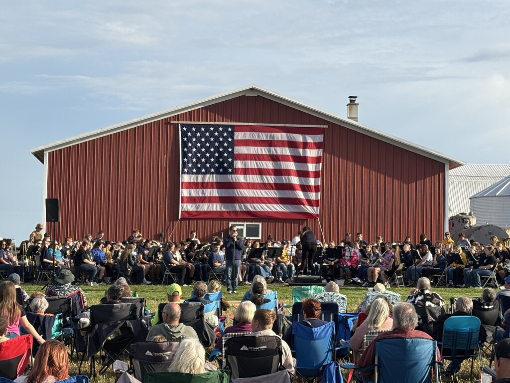 A full band sits in front of a barn with a large american flag on it