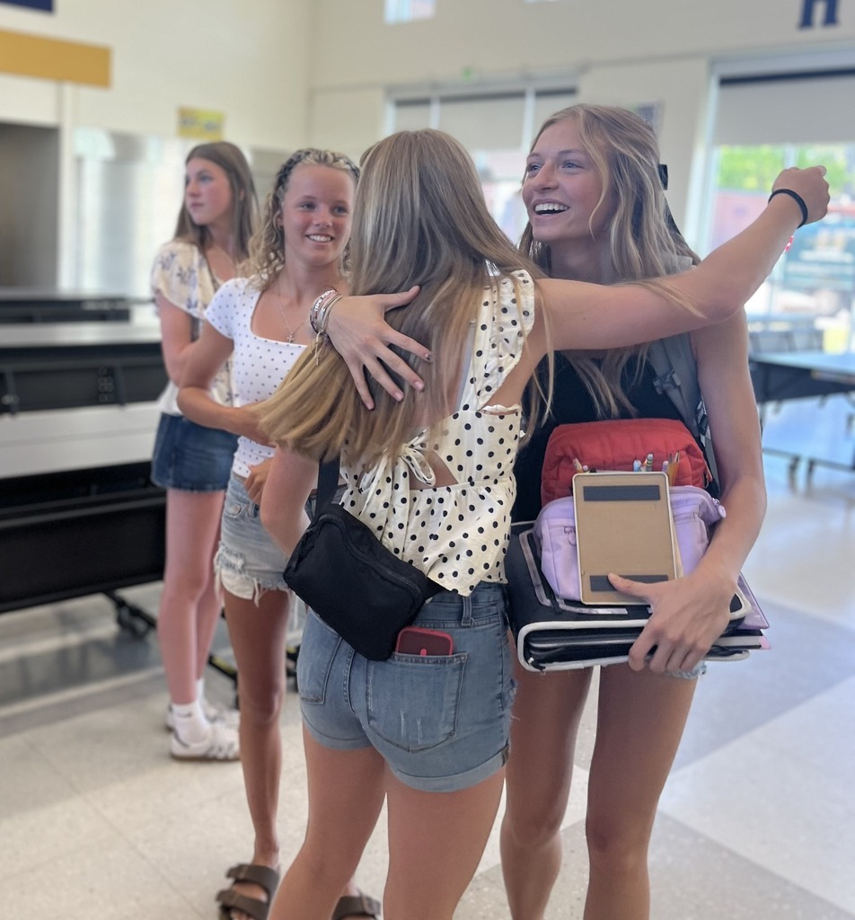 Two young female students hug with smiles on their faces. Two more students are in the background watching on.