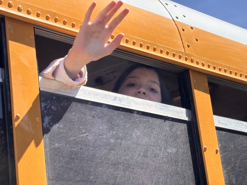 A young girl peaks out from a partially open bus window and waves goodbye