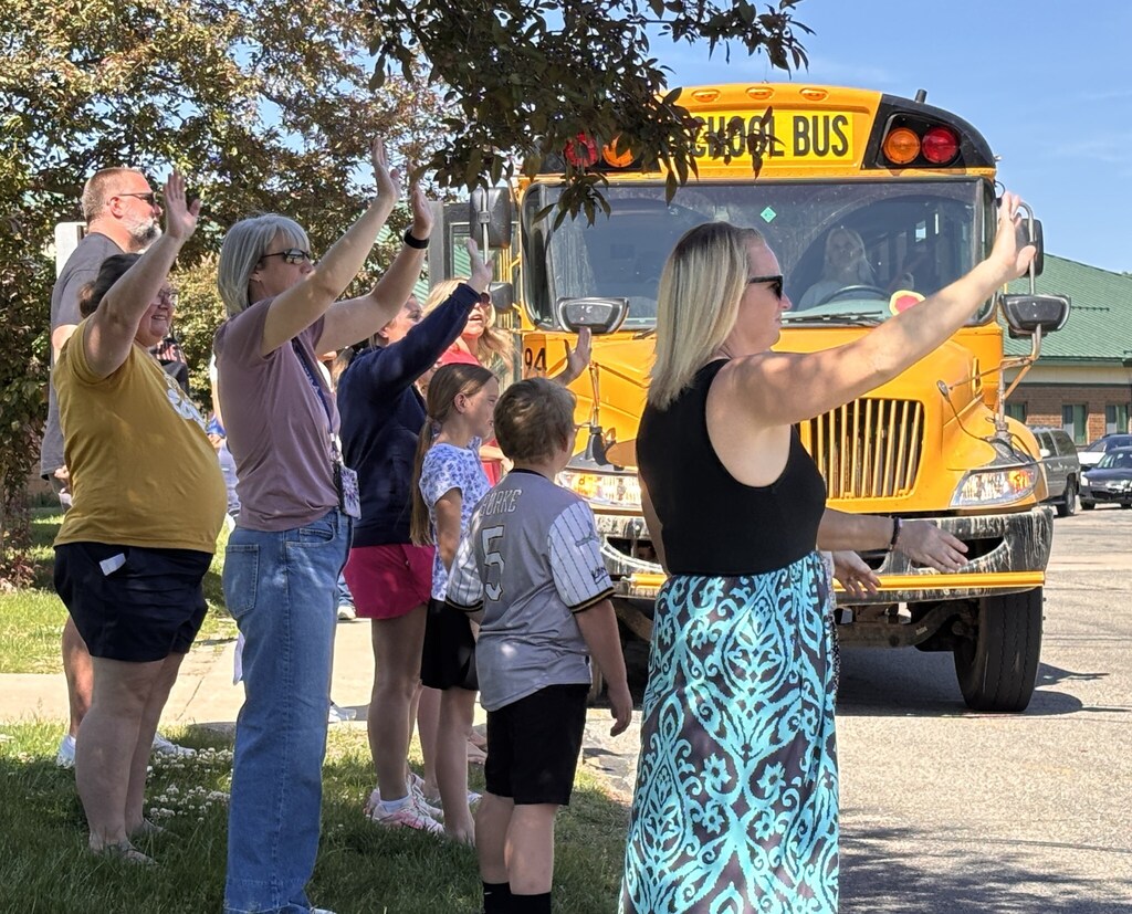Teachers line up by a parking lot and wave at departing students