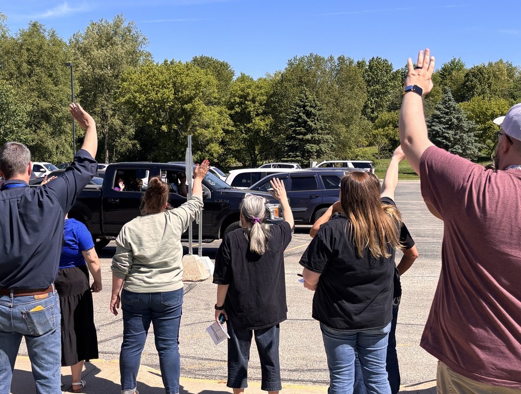 Teachers line up by a parking lot and wave at departing students