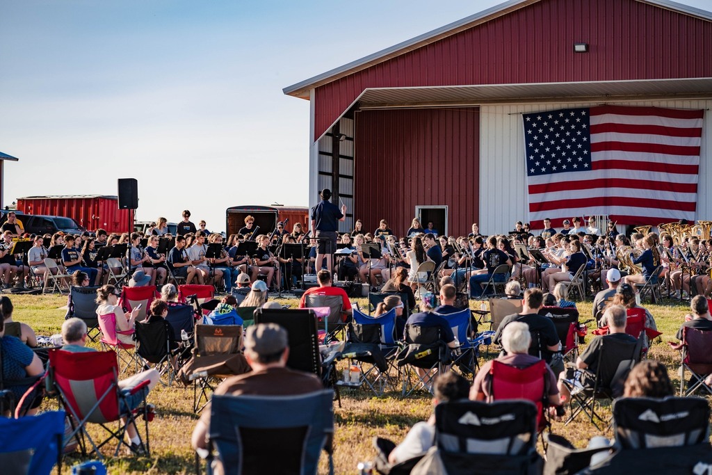 Multiple people are seated in lawn chairs as they watch a band perform in front of a barn with a large American flag.