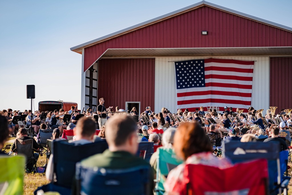Multiple people are seated in lawn chairs as they watch a band perform in front of a barn with a large American flag.