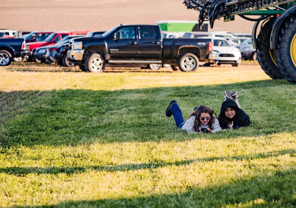 Two girls are laying in the grass with vehicles parked behind them
