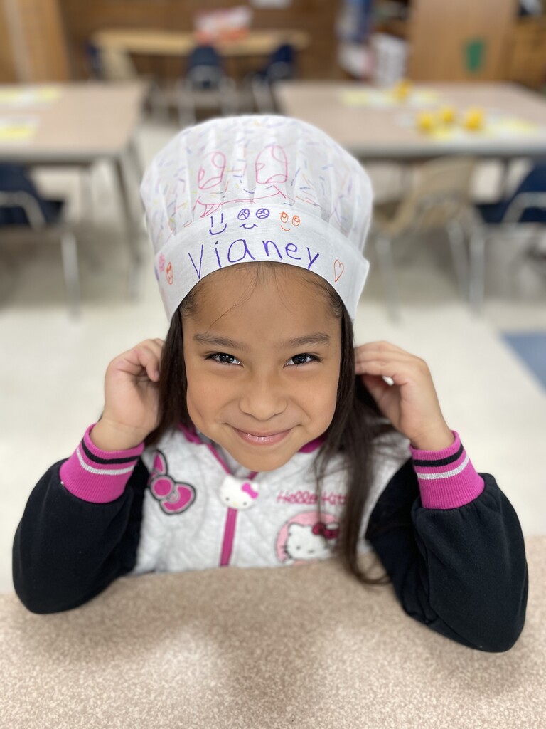 A girl smiles as she hold on to a chef hat that is perched on her head