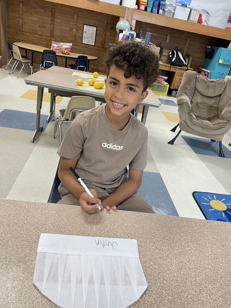 A boy sits behind a desk holding a marker as he writes his name on the brim of a homemade chef hat