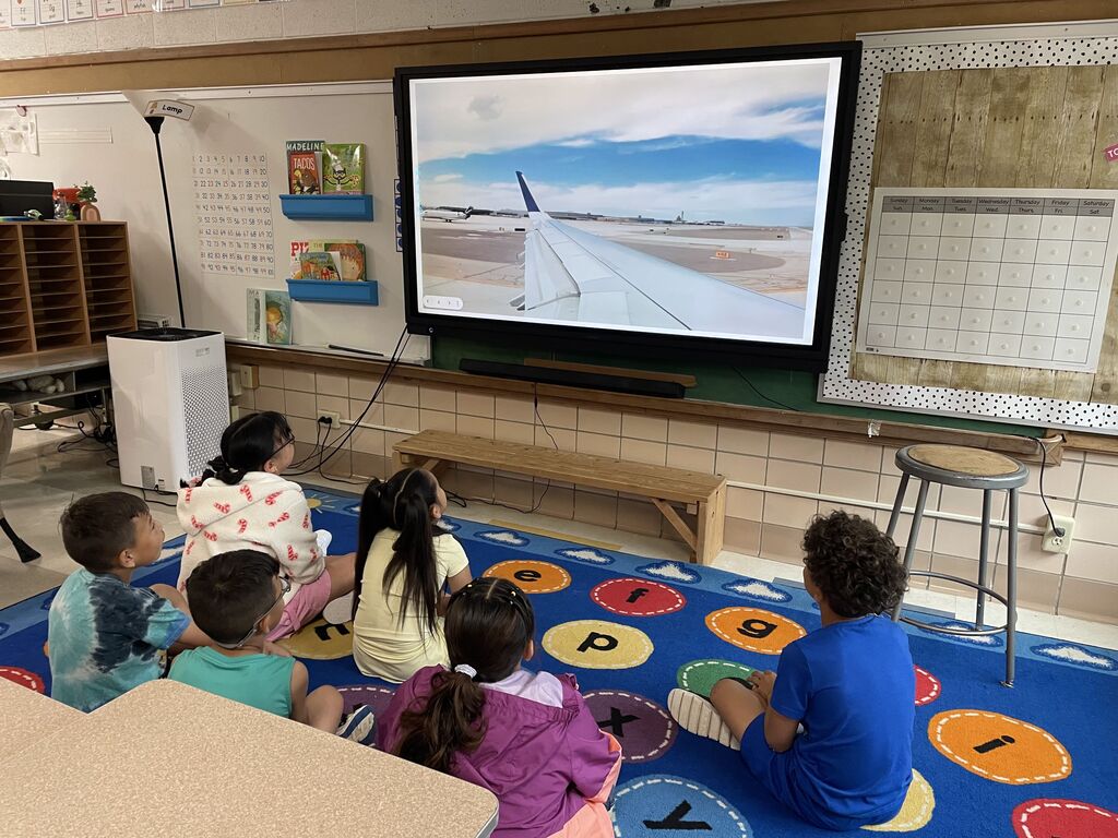 Children sit on the ground as they watch a movie screen that is displaying images of an airplane