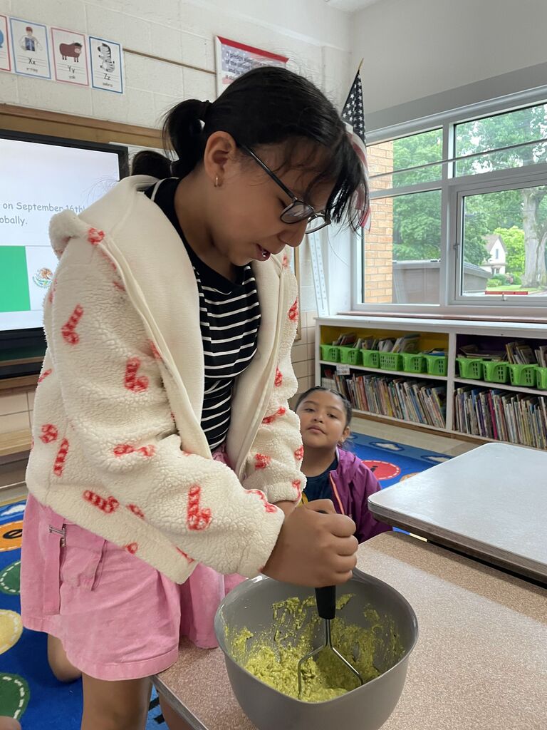 A girl wearing a fleece jacket is standing and crushing up a green substance in a mixing bowl