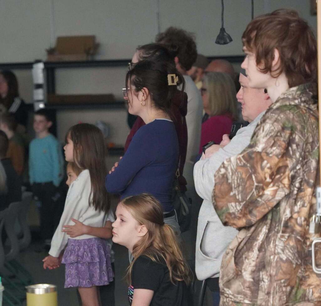 A group of parents and kids stand watching something that is out of the frame.