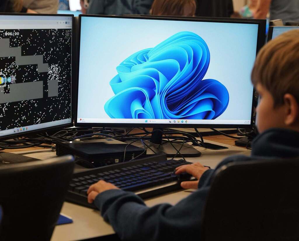 A boy sits in front of two side-by-side computer screens, one with a swirling blue design on the screen,