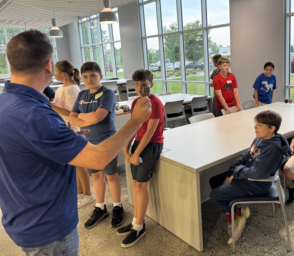 A man in the forefront holds up a metal object while a group of kids in the background look on.