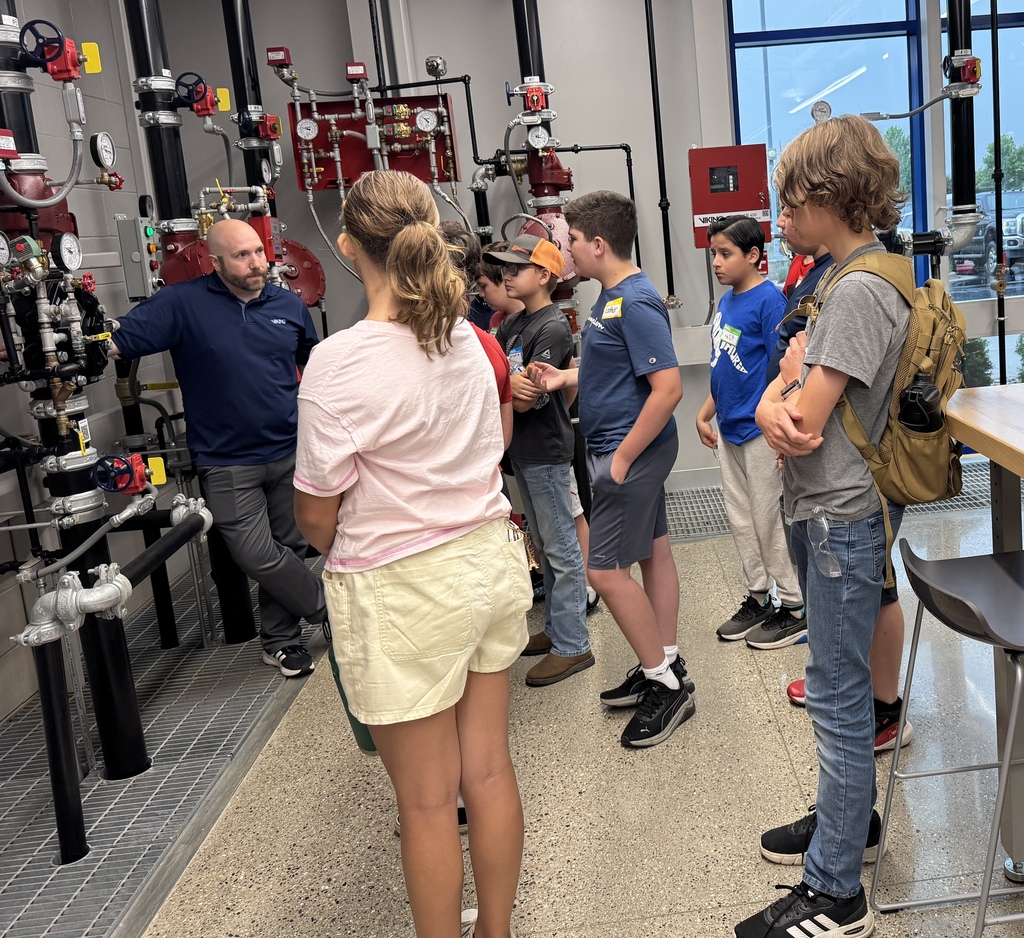 A man in a blue shirt faces a group of students as he displays a series of pipes that are fixed to the wall.