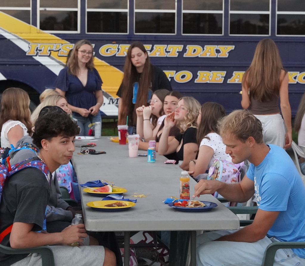 Students are seated on both sides of a long table eating breakfast and talking to each other.