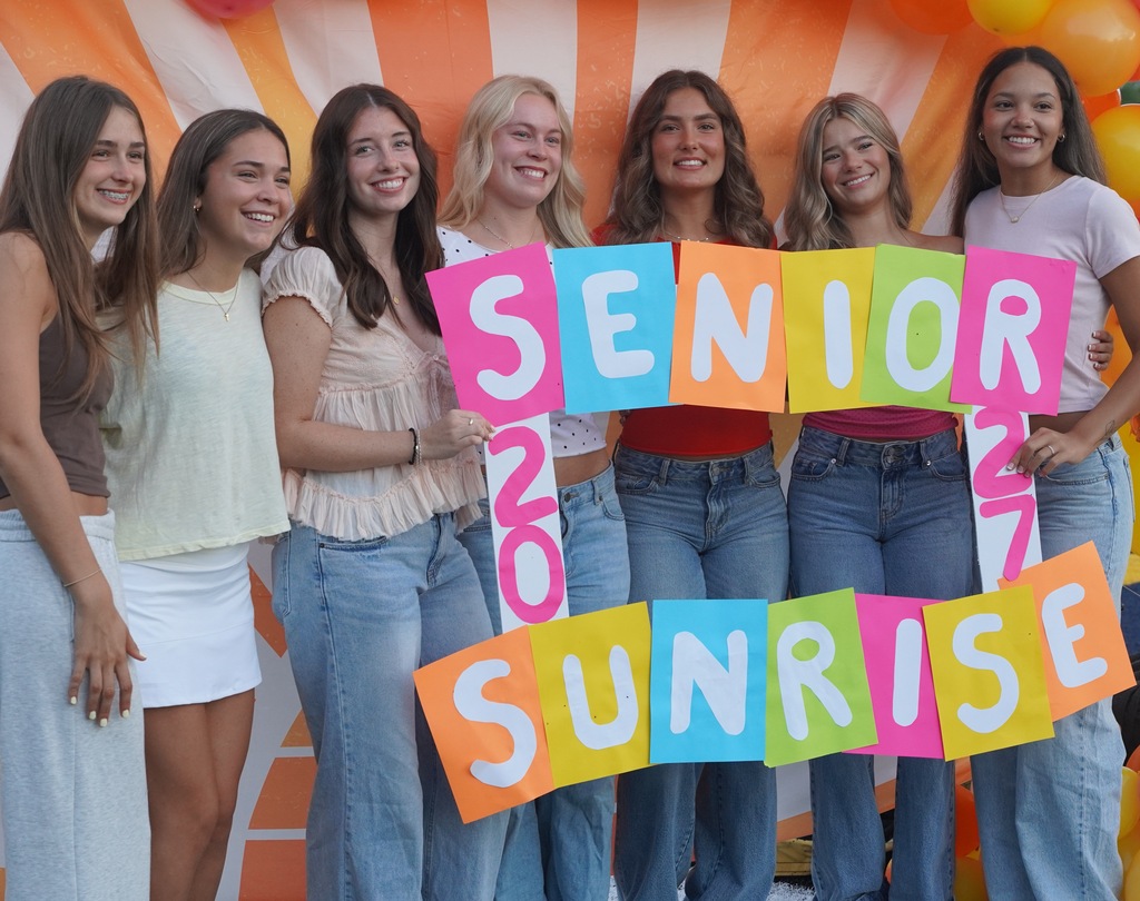 Seven female students pose for a picture with a colorful background behind them. They're holding a sign that says "Senior Sunrise"