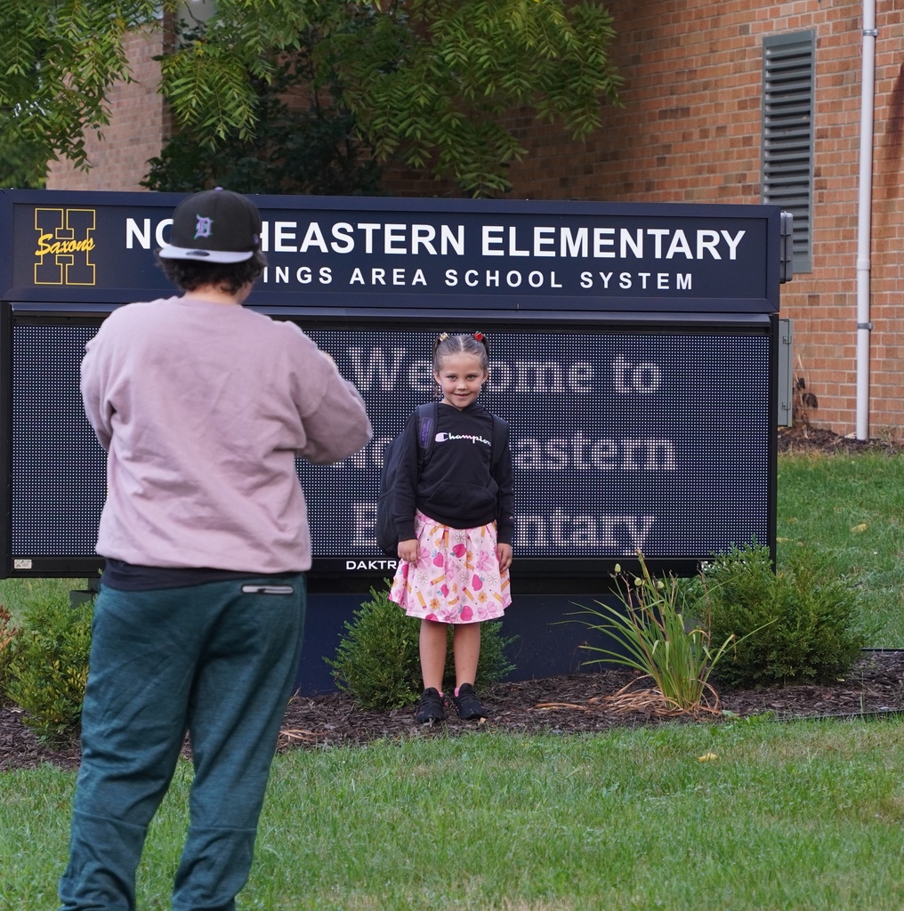 A girl stands in front of her school sign with a smile on her face while her mom takes a photo.