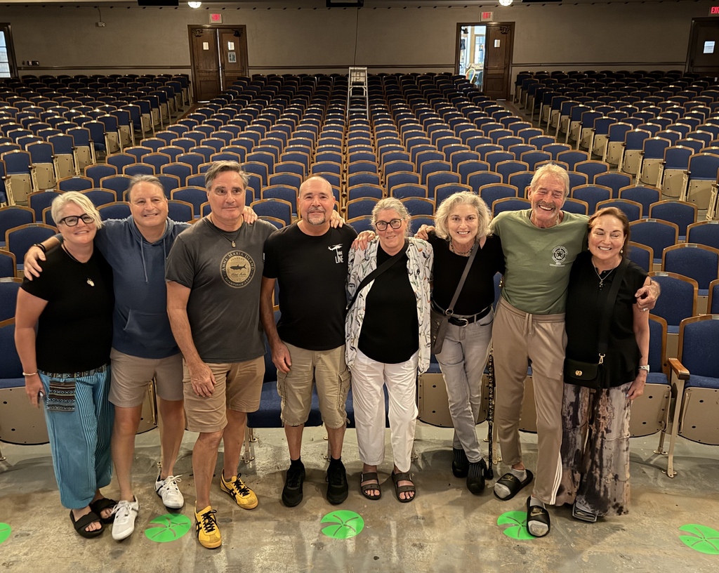 A group of middle-aged siblings stand in an auditorium with chairs behind them cposing for the camera.