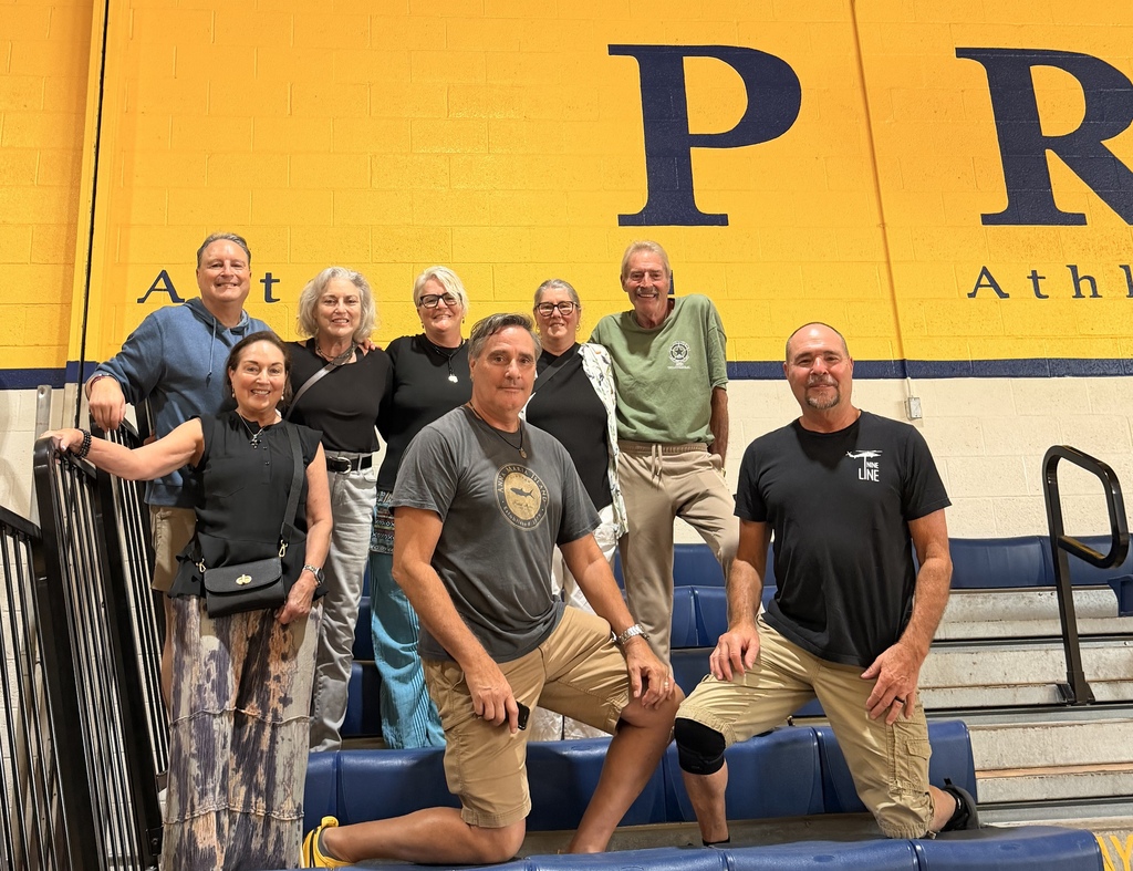 A group of middle-aged siblings stand on gym bleachers together with a bright yellow wall behind them posing for the camera.