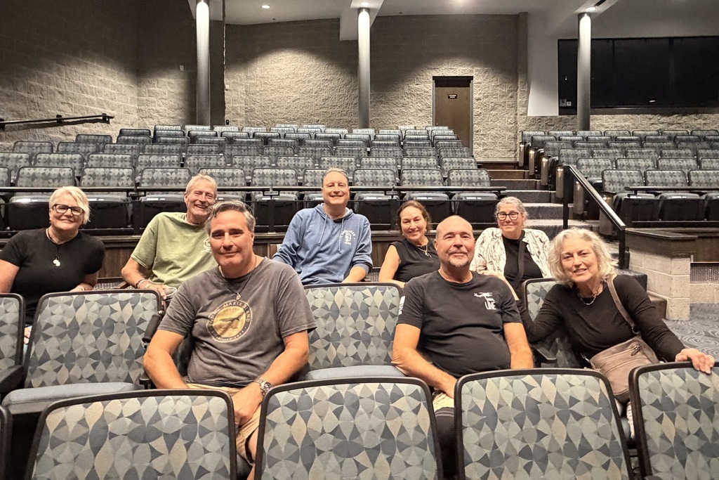 A group of middle-aged siblings sit in auditorium chairs posing for the camera.