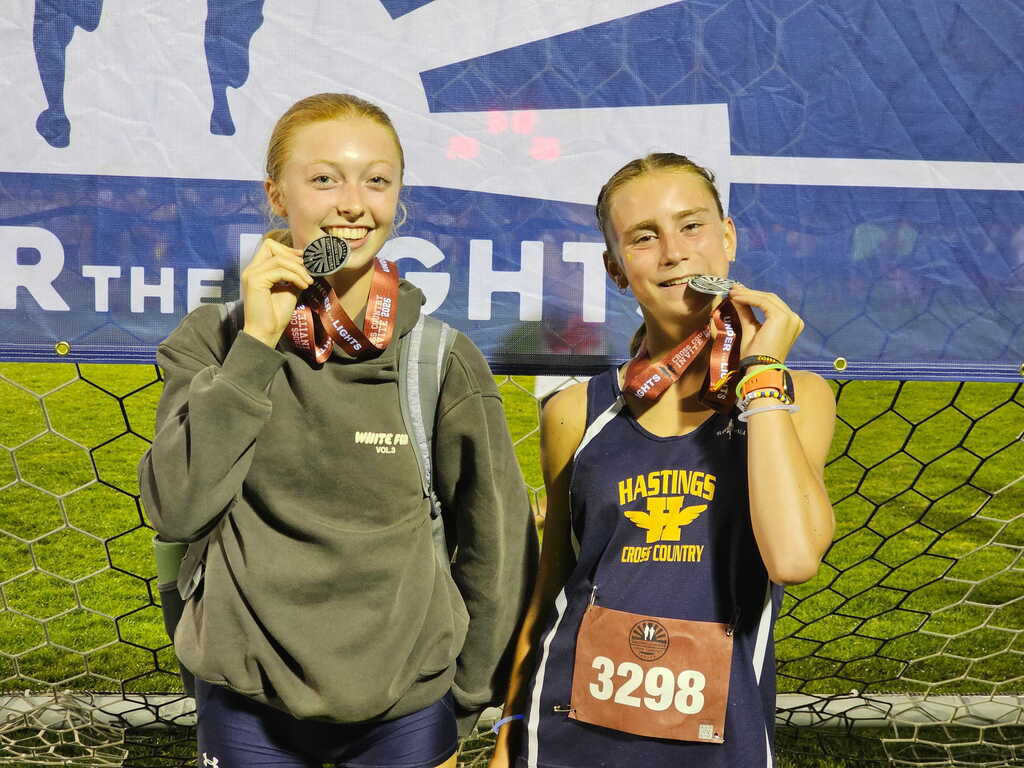 Two young females are standing in front of a blue banner with medals around their necks. They are biting the medallion of it as they pose.