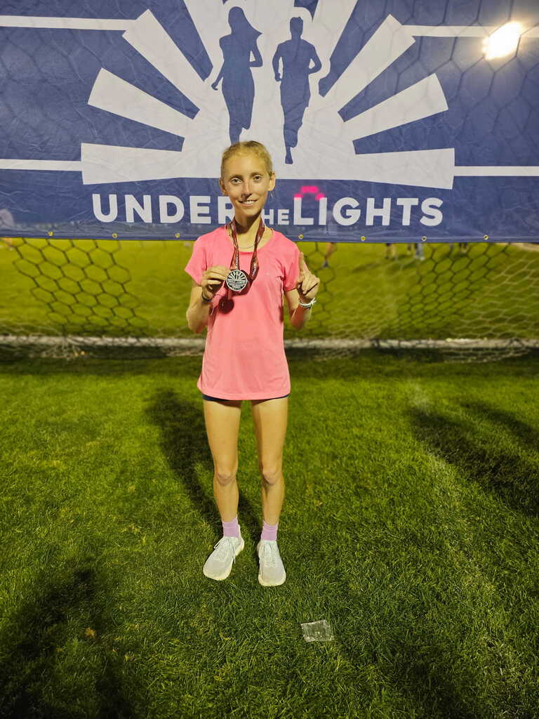 A girl in a pink shirt stands in front of a blue banner with a medal around her neck and holding up one finger.
