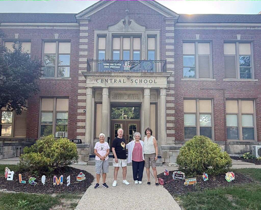 Three older individuals stand in front of a brick school building that reads Central School on the front.