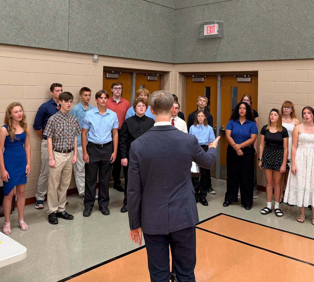 A choir director stands with his back to the camera as he directs a large group of boys and girls in front of him.