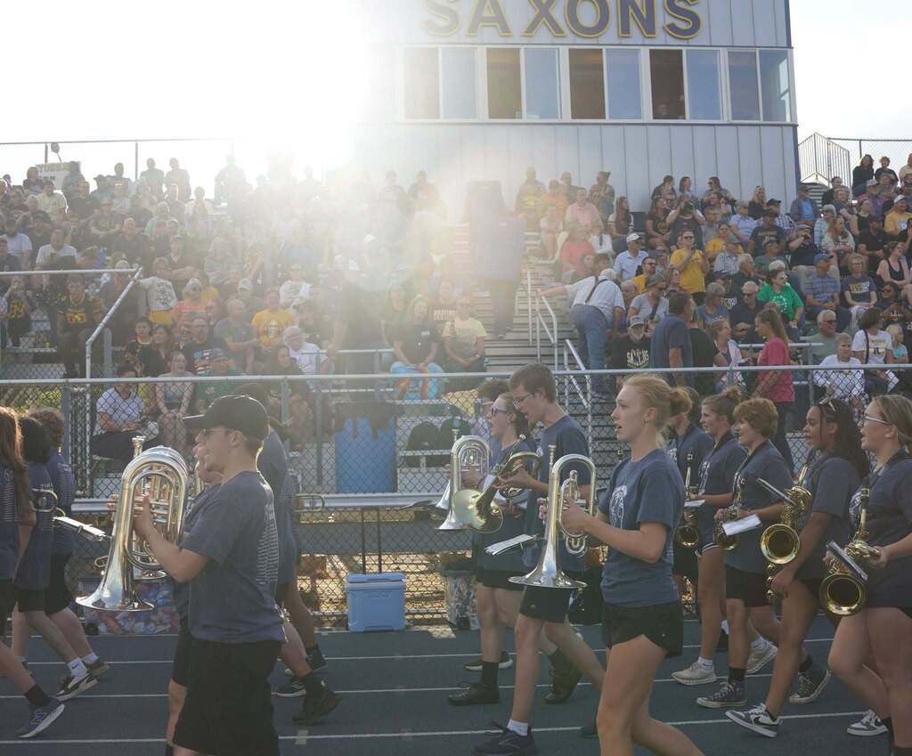 A marching band walks along the track with a crowded stands of people in the background.