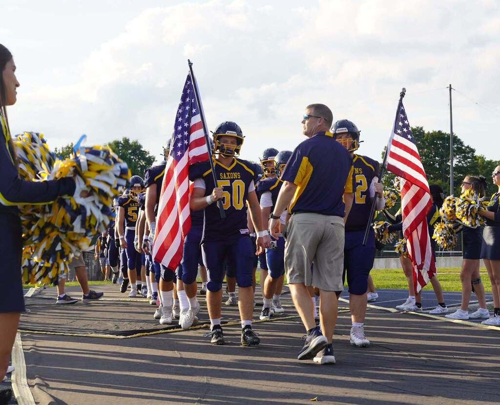 Two football players in dark blue uniforms holding American flags prepare to run their team out on to the field.