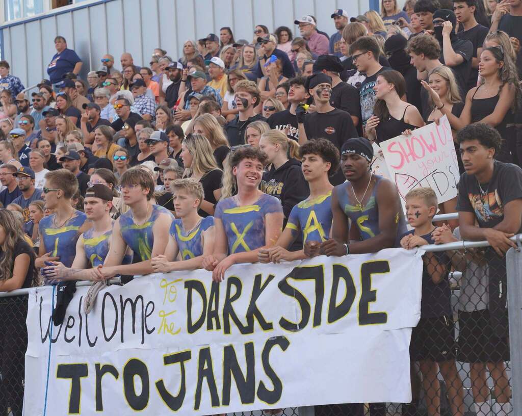 A large group of high school kids sit a section of bleachers. They draped a banner over the edge that reads "Welcome to the Dark Side Trojans"