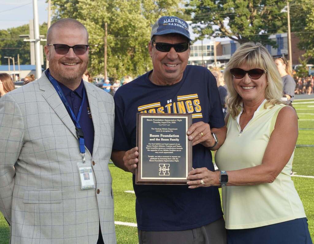 A man with a suit stands to the left of a couple, who are holding a plaque. All three are smiling for the camera. In the background you can see they are standing on a football field.