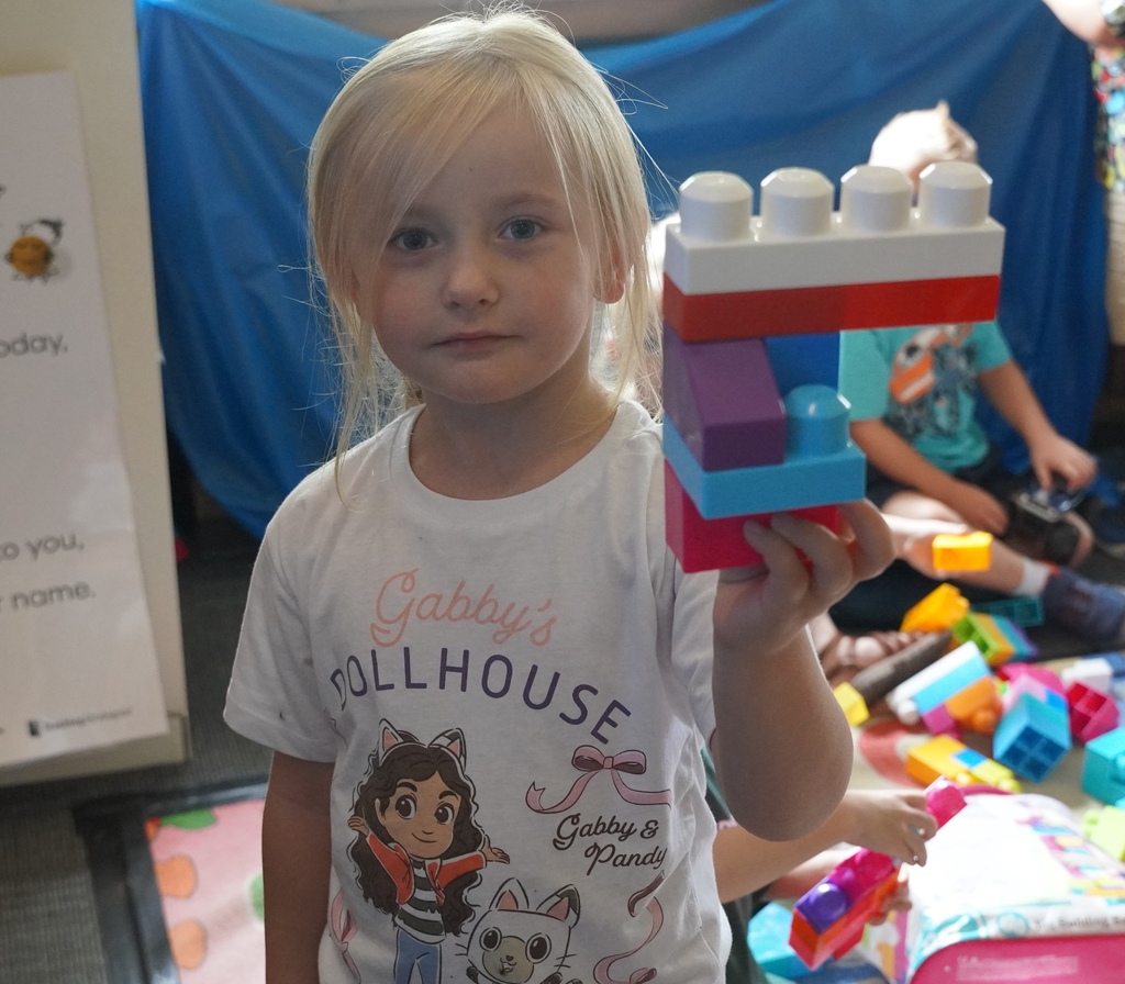 A young girl with blonde hair holds up a stack of jumbo legos that she assembled.
