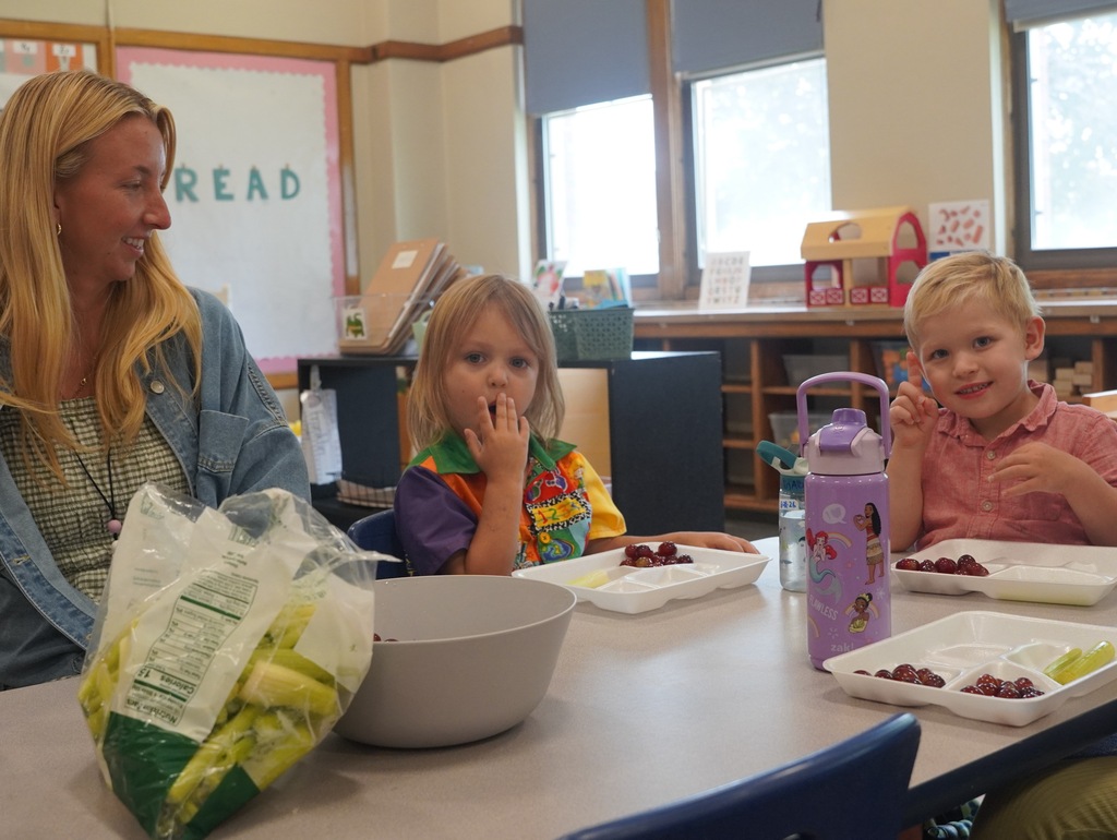 A teacher on the far left watches on as two little kids look into the camera. A girl is putting some food in her mouth while the boy gives a slight wave.