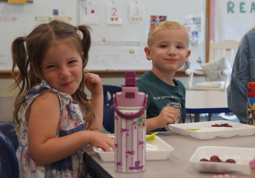 Two children sit at a table eating lunch. The girl on the left, with pigtails, smiles at the camera with a boy next to her who is also smiling.