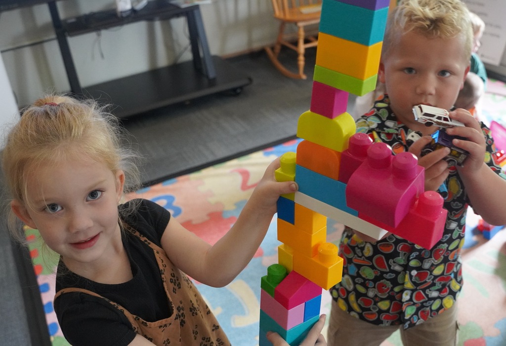 Two young kids show off their toys to the camera. The girl shows a stack of jumbo legos she assembled and the boy holds up a small toy car.