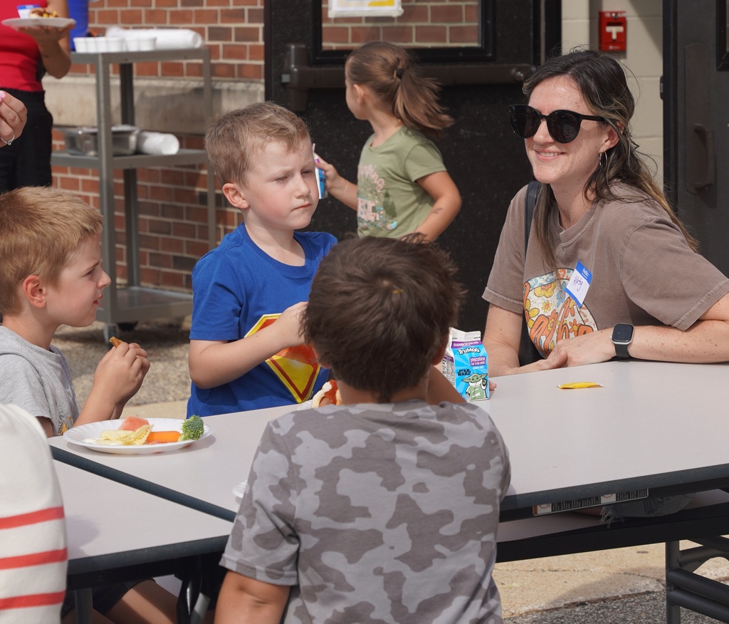 A woman sits sideways at an outdoor table facing three kids who are eating off styrofoam plates.