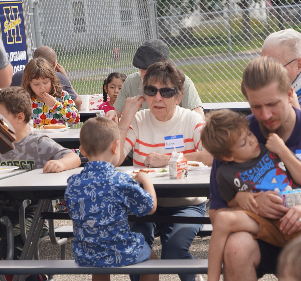 A woman with sunglasses sits at a table outdoors, facing a young child whose back is to the camera.