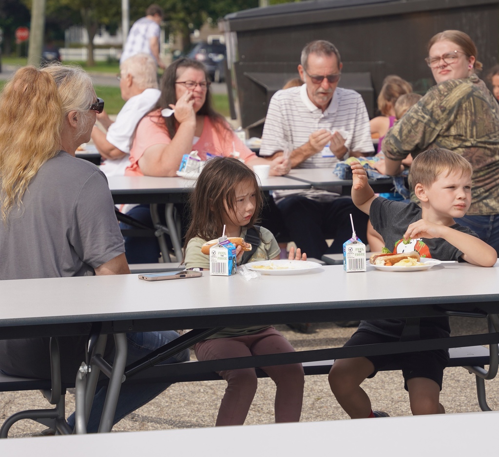 Two young kids, a girl and boy, sit at a table outdoors with others seated in the background.
