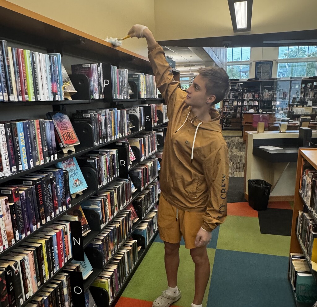 A male high school student with a tan hoodie on reaches to the top of a book shelf to dust it.