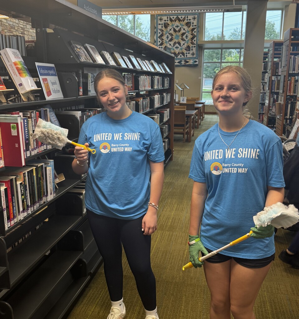 Two female students hold dusters as they smile at the camera. They're both wearing the same blue shirt that says "United We Shine" on it.
