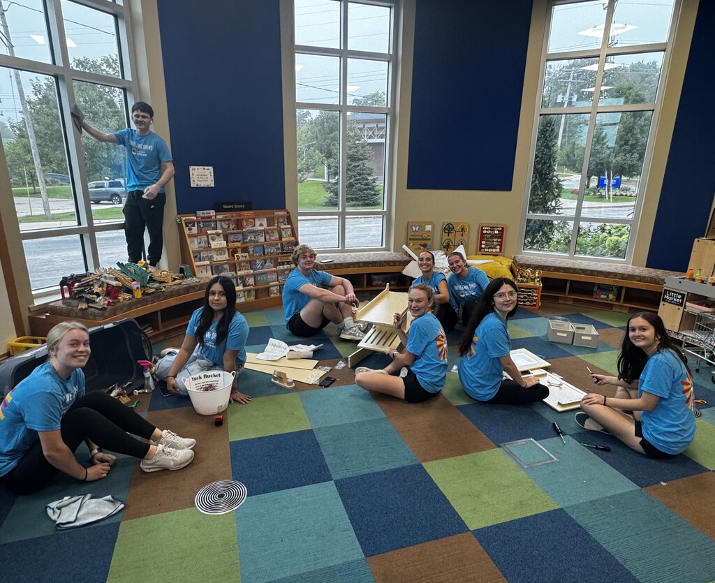 A group of high school students with blue shirts on pause from their work and smile at the camera. They are sitting on a carpted floor organizing items.
