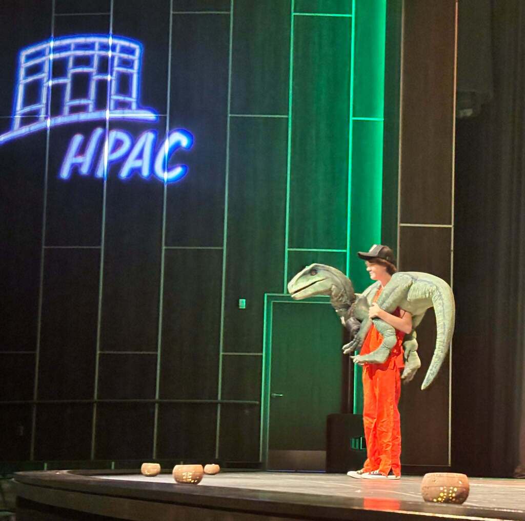 A young person in a jumpsuit holds a dinosaur around their neck on a stage.