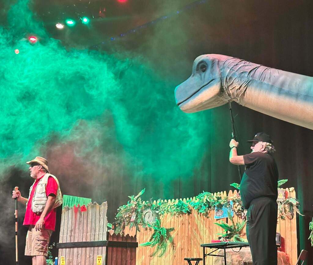 Fog hovers over a stage as a man dressed in safari gear looks off camera and a large dinosaur head peaks out from the right of the frame.
