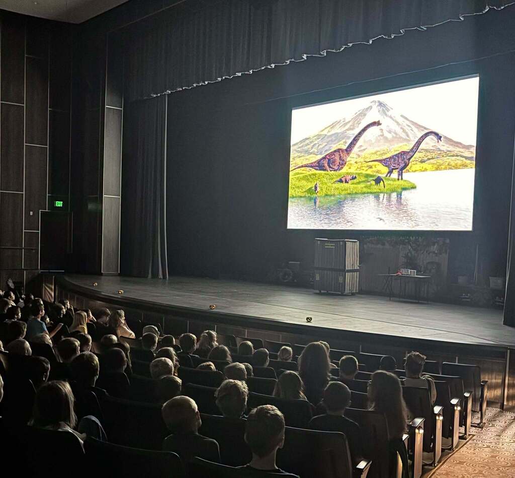 Students fill auditorium seats as they gaze up at the stage where a movie screen is showing photos of dinosaurs.